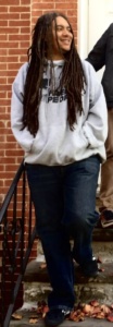 The image is a picture of a light-skin Black man with long locs smiling with his face turned towards the right. He is wearing jeans and a gray long-sleeved sweatshirt that reads, "Free Minds Free People" in bold black letters 